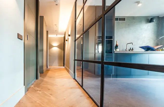 Een strakke, lichte hallway met warme houten naadloze vloer, witte wanden en elegante zwarte glazen scheidingswanden. Rechts zicht op een minimalistisch badkamer- of wasruimte-gedeelte met subtiele verlichting en designer accessoires.
