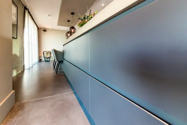 Een moderne keukenbar met een elegant gestyled interieur. De naadloze wand in zachte blauwgrijze tint loopt naadloos door, afgewerkt met subtiele streeplijn-detail. Heldere belichting en opgeruimde styling tonen de perfecte afwerking.