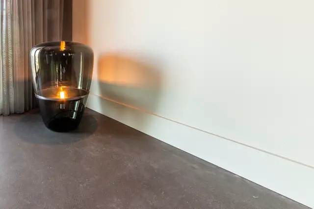 Een minimalistische woonruimte met naadloze vloer in warm grijs, scherpe lijnvoering langs de wand, en een elegant zwarte glazen designlamp met gouden gloed. De ruimte ademt rust en onderstatement uit.