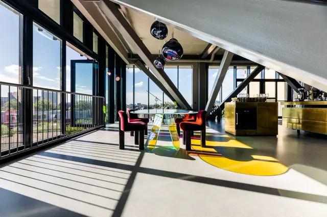 Ruime, licht inundated loft met naadloze vloer in natuurtinten en geel-groene accenten. Industriële architectuur met grote raampartijen, zwarte staalconstructie en designmeubels creëren een stijlvolle, openhartige leefruimte.