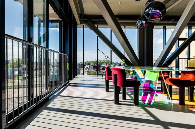 Ruim, licht terras met gestreepte naadloze vloer en zwarte constructieve elementen. Kleurrijke designmeubels en helder uitzicht over water en groen.