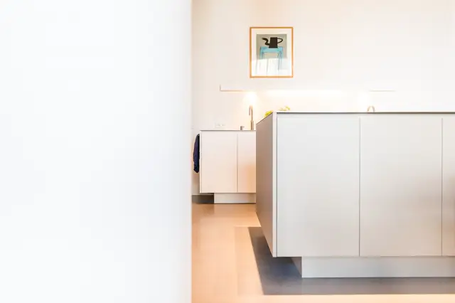 Strakke, lichte keukenruimte met witte inbouwkasten en naadloze wand. Zacht warm licht benadrukt de schone lijnen en rustige kleurpalet.
