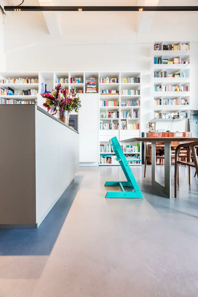 Open woonkamer met ingebouwde boekenkasten en een continue naadloze vloer in subtiele tinten. De ruimte is licht, minimalistisch ingericht met natuurlijke belichting en een elegant afgewerkt oppervlak.