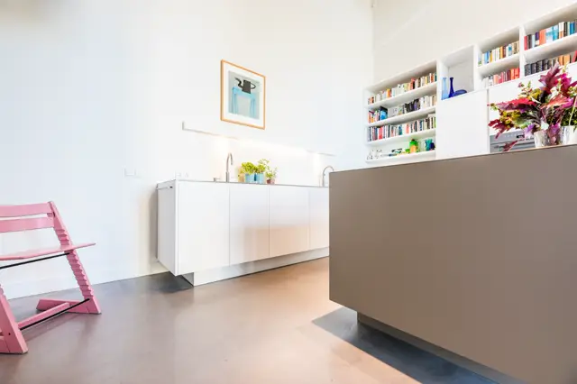 Een strak ingerichte woonkamer met gladde, grijsbruine naadloze vloer en wand. Witte inbouwkasten, wit keukenblok en taupe partitie creëren een serene, georganiseerde sfeer met speelse roze accentstoel.
