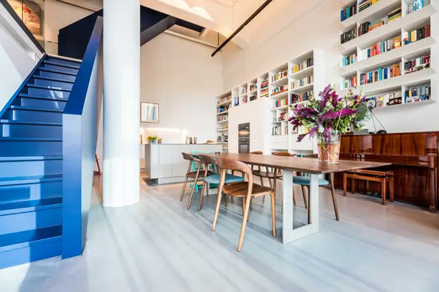 Ruim, licht interieur met open keuken en eetkamer. Een naadloze vloer in warm grijs loopt over twee niveaus en creëert een samenhangend, strak beeld. Witte wanden, blauwe trap, houten meubels en boekenkast.