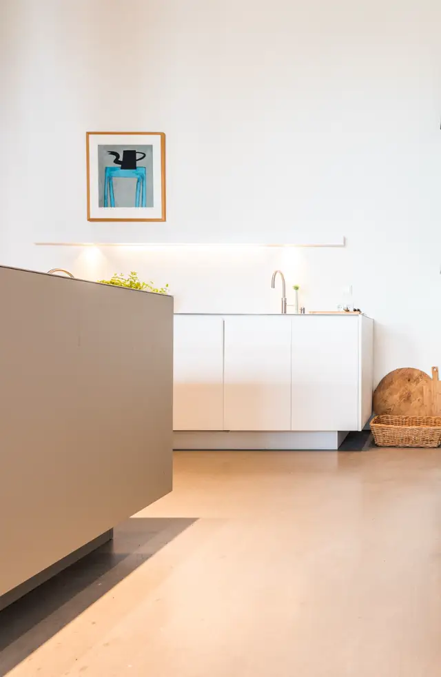 Serene keukenruimte met witte kasten, roestvrij stalen kraan en warme naadloze vloer. Licht stroomt natuurlijk naar binnen; wanddecoratie en planten accentueren de strakke, luxe esthetiek.