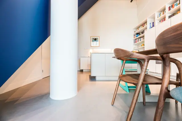 Strak ingerichte open woonkamer met heldere naadloze vloer, minimalistische witte meubels, houten designstoelen en een opvallend blauw accent aan de muur. Natuurlijk daglicht creëert een rustige, geordende sfeer.