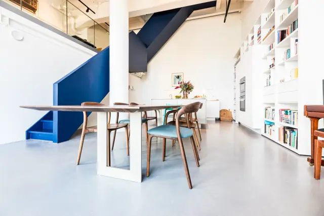 Ruim, licht interieur met naadloze vloer in zacht grijs-blauw. Strakke witte wanden, geometrische blauwe trappartij en open keukenlijn creëren een minimalistische, eigentijdse uitstraling.