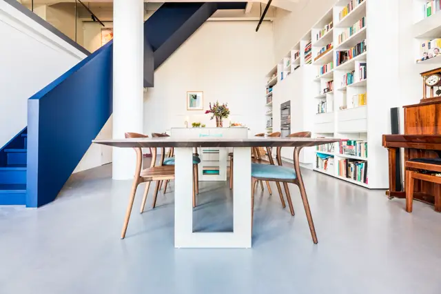 Ruime, licht ingerichte woon- en eetkamer met een hellend plafond, blauwe trap, witte inbouwkasten en een elegante eettafel. De naadloze vloer loopt naadloos door de hele ruimte en creëert rust en continuïteit.