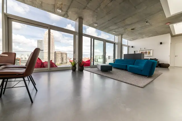 Ruime loft met gladde, grijze naadloze vloer die de hele open ruimte verbindt. Groot glasfront met panoramisch uitzicht op de skyline, minimalistisch ingericht met blauwe modulaire bank en designmeubels.