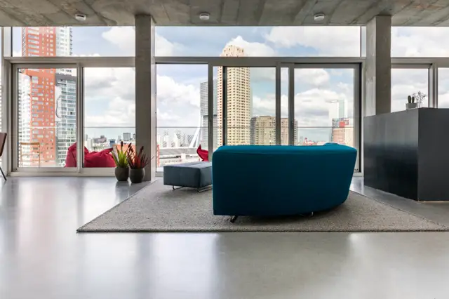Ruim, licht penthouse met naadloze grijze vloer, betonnen plafond en panoramische ramen met uitzicht op de stad. Minimalistisch ingericht met blauwe fauteuil en geometrische vormen.