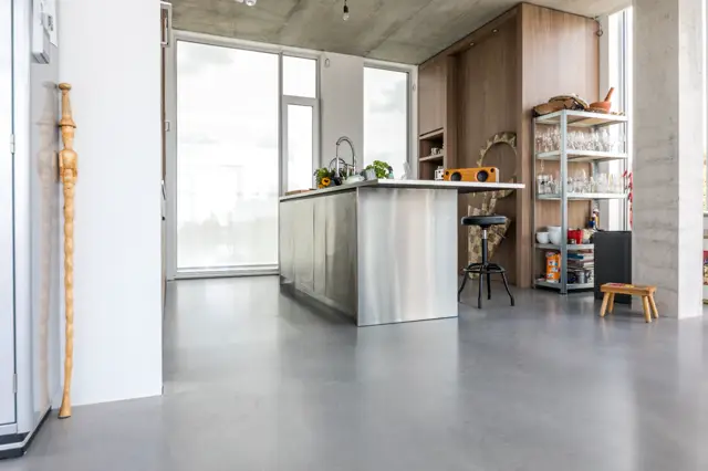 Minimalistisch ingerichte loft met een doorlopende naadloze vloer in grijs beton. Het ruime, lichte interieur combineert industriële elementen met warme houten accenten en moderne keukenuitvoering.