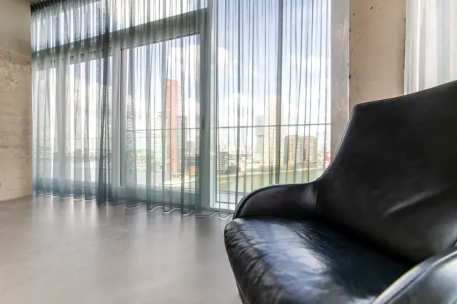 Ruime, lichte woonruimte met grote raampartijen en transparante gordijnen. De naadloze vloer loopt naadloos door naar het zitgedeelte met design stoel. Strakke lijnen en minimalistisch interieur.