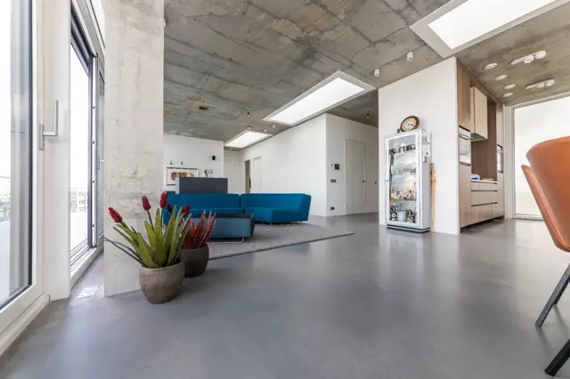 Spacieuze open loftruimte met uniforme naadloze vloer, industriële betonplafond, grote ramen en minimalistisch interieur met blauwe zithoek en houten accenten.