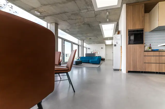 Ruim, licht openplan interieur met naadloze vloer in grijs. Industriële betonnen plafondstructuur en grote raampartijen creëren een modern loft-karakter met houten keukenkasten en design meubels.