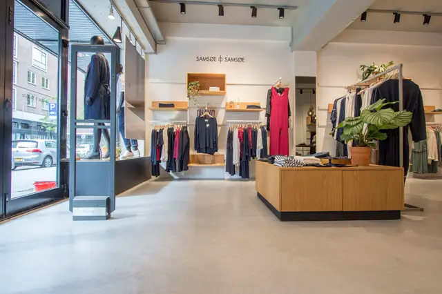 Minimalistisch ingerichte kledingboetiek met een strakke, lichte naadloze vloer die het hele pand doorloopt. Helder daglicht, houtaccenten en opgeruimde merchandising creëren een luxe winkelervaring.