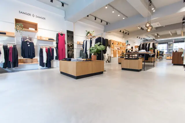 Ruime, lichte winkelruimte met een naadloze vloer in licht grijs. De ruimte is strak ingericht met houten display-elementen, witte wanden en professionele verlichting. Perfecte zichtlijnen en volledige opgeruimdheid.