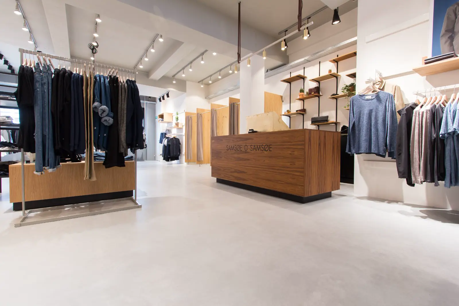 Een strakke retail ruimte met naadloze lichte vloer, wit plafond met geïntegreerde verlichting en houten display elements. Schone lijnen, opgeruimde presentatie van kleding op hangers en houten toonbank.