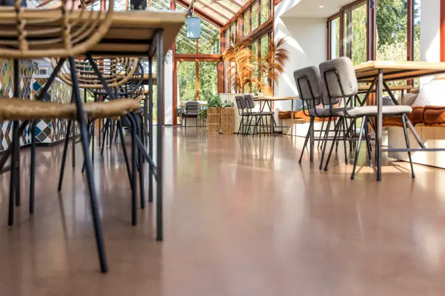Ruime, lichte loungeruimte met een egale naadloze vloer in warm grijs. Grote raampartijen bieden uitzicht op groen, moderne stoelen en natuurlijke materialen creëren een rustgevende sfeer.