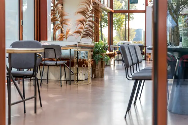 Ruime, licht ingestelde bedrijfsruimte met een naadloze vloer in warm grijs. Strakke inrichting met moderne barkrukken, plantendecoratie en grote ramen die natuurlijk licht binnenlaten.