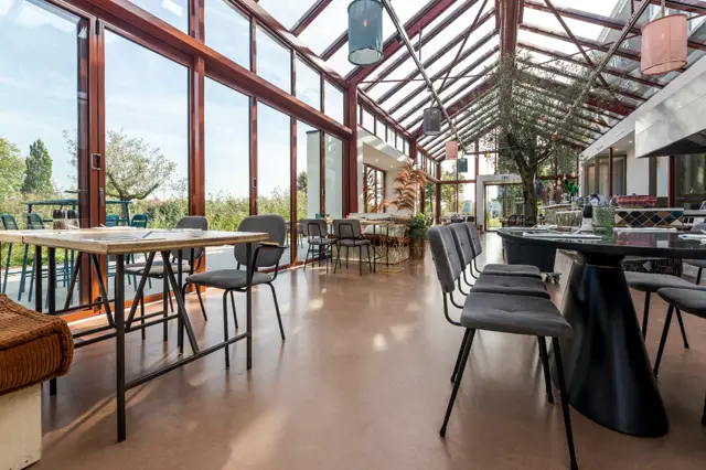 Ruim, licht restaurant- of eventruimte met glazen overkapping en stalen constructie. De naadloze vloer in warm grijs loopt ononderbroken door, wat de airy openheid versterkt. Minimalistisch ingericht met designmeubelen.
