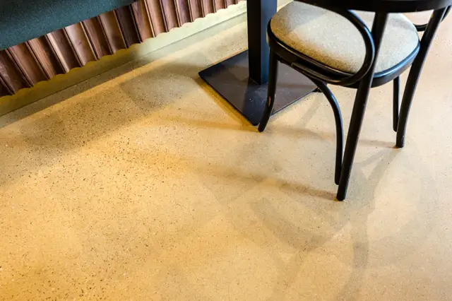 Een elegant ingerichte kantine met een warmgetinte naadloze vloer, zwarte stoelen met naturelle zitting en houten accenten. De vloer loopt naadloos door en creëert een rustige, geïntegreerde uitstraling.