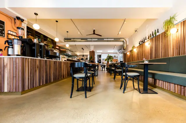Spacious, lichtinval cafetaria met een warme, uniforme naadloze vloer die zich elegant uitstrekt over het gehele interieur. De vloer creëert een rustgevoel en ondersteunt het strakke, minimalistische design met houten accenten en groene bouwlijn.