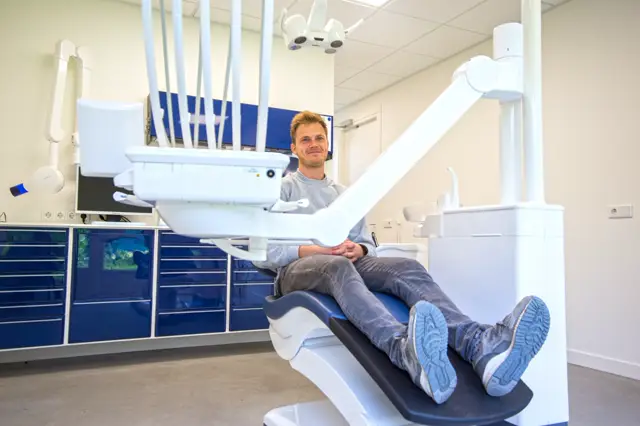 Een schone, lichte tandartskliniek met strakke witte apparatuur, donkerblauwe kasten en een neutrale naadloze vloer. De ruimte oogt professioneel, hygiënisch en georganiseerd.