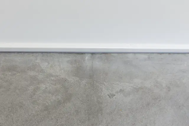 Detail van een naadloze wand in warme grijstoon met subtiele textuur, afgezet met een strakke witte horizontale lijnen. De afwerking toont een rustieke, natuurlijke uitstraling met verfijnde kwaliteit.
