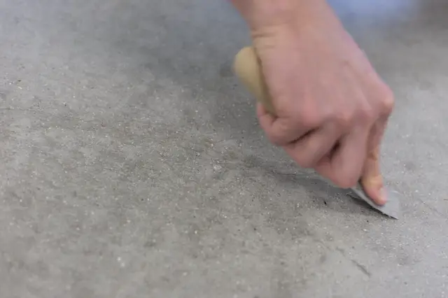 Close-up van een hand die de perfecte afwerking van een naadloze vloer controleert. De vloer vertoont een subtiele textuur met fijne korrelstructuur en neutrale grijs-beige toon.