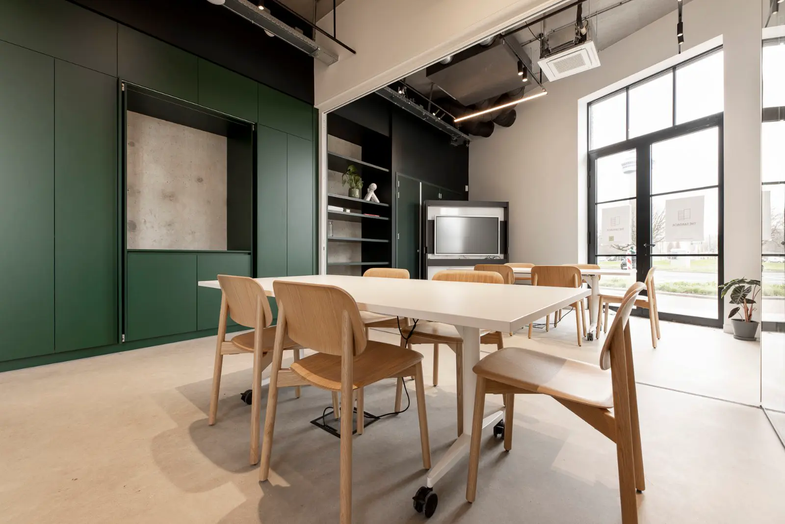 Strakke bedrijfsruimte met lichte naadloze vloer, groen-zwarte wandkasten en natuurlijk licht. Moderne, opgeruimde werkplek met houten meubels en grote ramen.