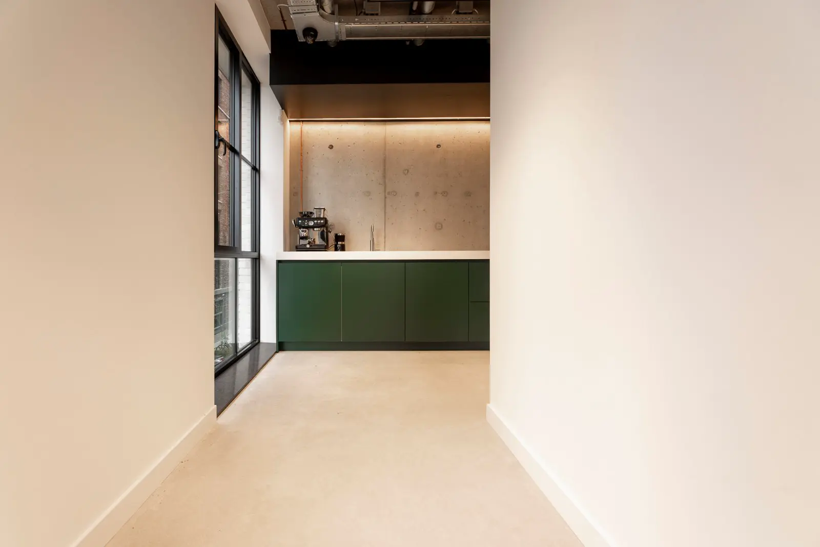 Strakke open keukenruimte met een helderbeige naadloze vloer, donkergroene kasten en een warm verlichte betonnen wand. Een architectonisch verfijnde opstelling met rechte lijnen en subtiele materiaalcontrasten.