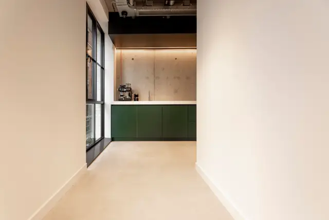 Strakke open keukenruimte met een helderbeige naadloze vloer, donkergroene kasten en een warm verlichte betonnen wand. Een architectonisch verfijnde opstelling met rechte lijnen en subtiele materiaalcontrasten.