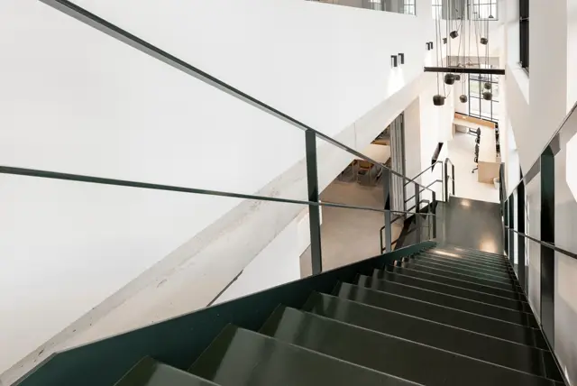 Een strakke trap met donkergroene treden leidt omhoog naar een minimalistisch interieur. Witte naadloze wanden, zwarte stalen leuning en hangende design-verlichting creëren een voornaam en open ruimtegevoel.