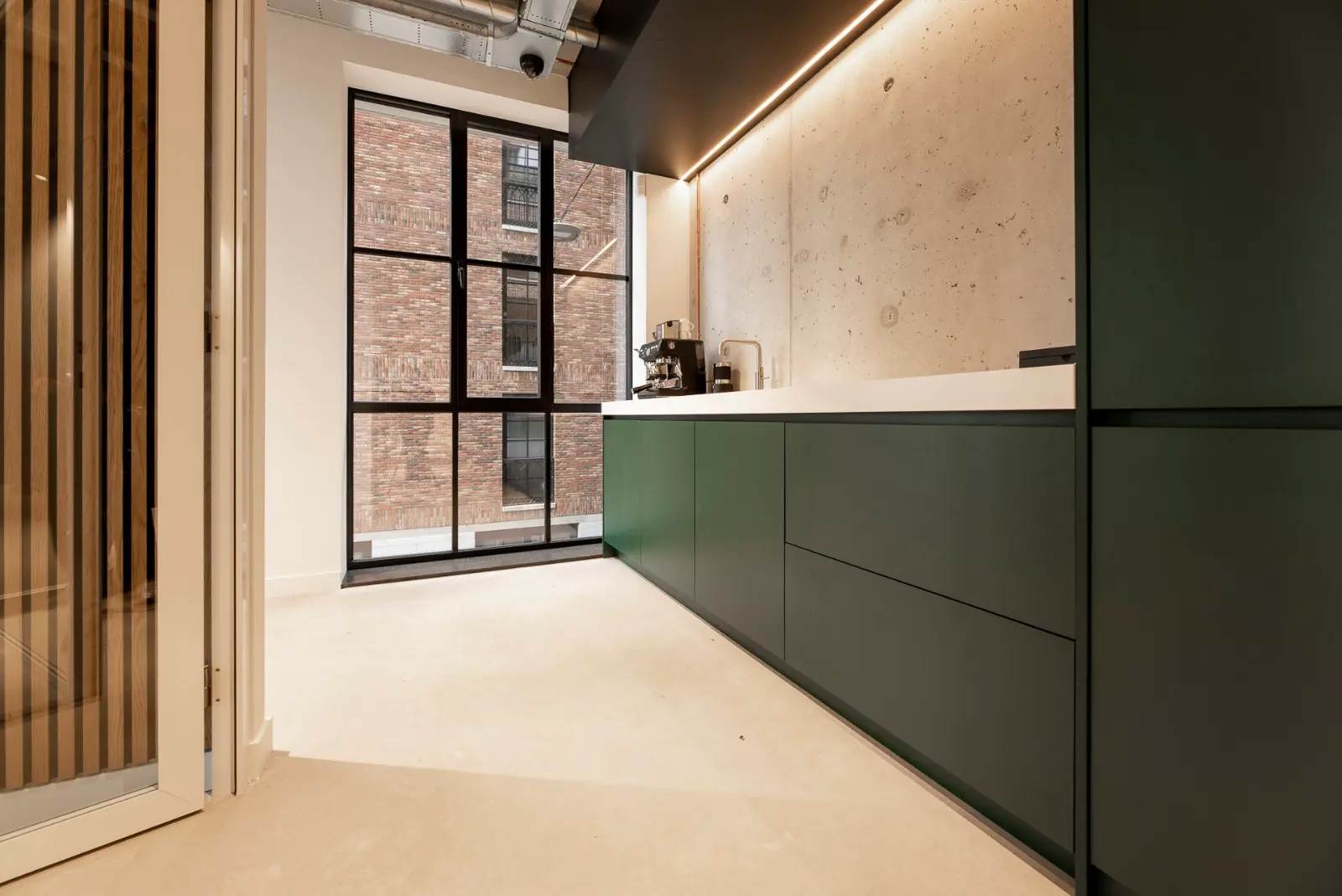 Een strakke, moderne keukenruimte met naadloze wand- en vloeroppervlakken in beige tinten. Groene kastelementen vormen een subtiel accentueel contrast tegen de lichte, geopende ruimte met rijke daglichtinval.