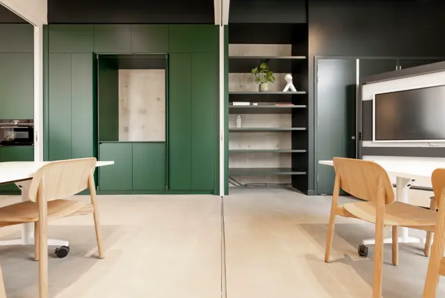 Een strak ontworpen open werkruimte met naadloze lichte vloer, groene kasten en donkere wanden. Natuurlijk licht, minimalistisch meubilair en opgeruimde styling creëren een rustieke, professionele sfeer.