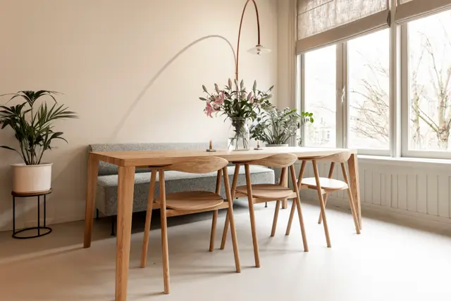 Serene eetkamer met natuurlijk licht, houten tafel en stoelen, grote ramen en subtiele plantendecoratie. De naadloze vloer creëert een schoon, doorlopend oppervlak dat de ruimte optisch vergroten.