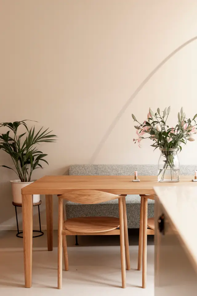 Een serene eetkamer met houten tafel en stoelen op een lichte, naadloze vloer. Zachte beige wanden met subtiele schaduwwerking creëren diepte; fresh bloemen in glas accentueren de rustieke elegantie.