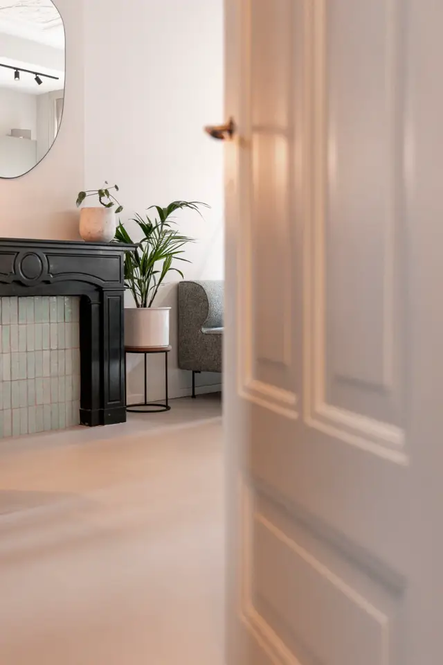 Een gerenoveerde hal met lichte, naadloze vloer en minimalistische inrichting. Zachte natuurlijke lichtinval door grote ramen, gestileerde details met zwarte haard en groene planten.