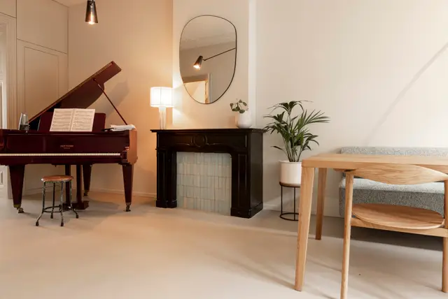 Een ruime, licht ingerichte woonkamer met een ononderbroken naadloze vloer in warm beige. De ruimte combineert klassieke meubelen (piano, haard) met minimalistisch design, ondersteund door subtiele verlichting en een opgeruimde, serene sfeer.