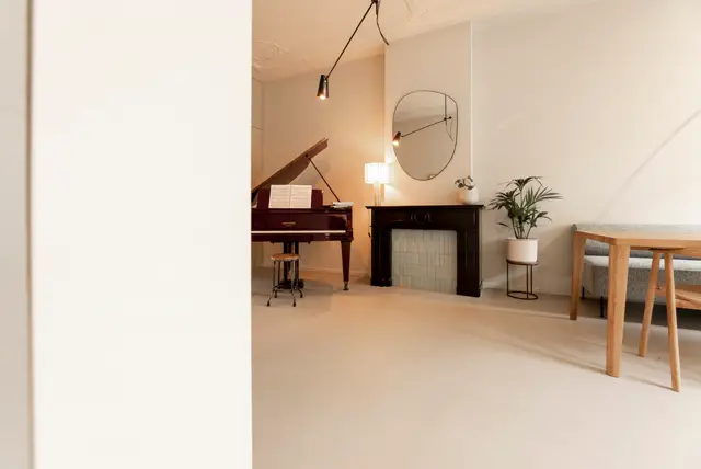 Serene, licht ingerichte ruimte met een continue naadloze vloer in warm beige. De open, minimalistische setting met vleugel, design meubels en natuurlijk licht creëert een rustige, premium sfeer.