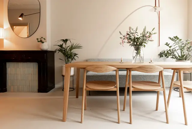 Een stijlvolle eettafel van hout met grijze stoelen staat op een naadloze vloer in warm beige. De wand erachter is schoon wit met subtiele bogenvormige architectonische details, versierd met verse bloemen en groene planten.