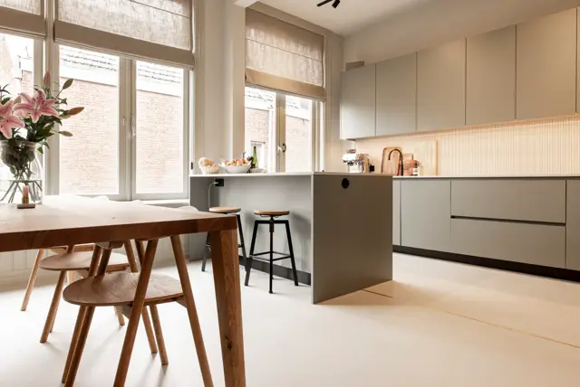 Minimalistisch ingerichte keuken met strakke grijsbeige kasten, naadloze crèmekleurige vloer en veel natuurlicht. Houten eettafel en gestileerde details creëren een rustig, geordend geheel.