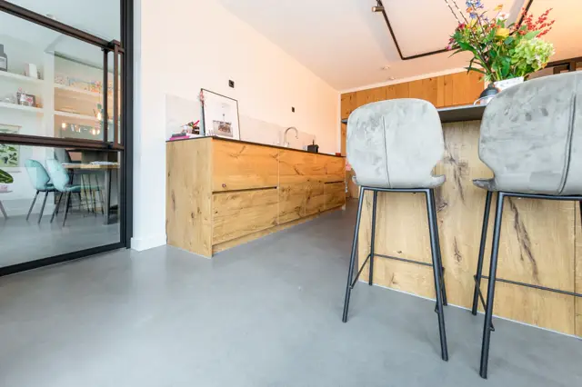 Strakke, lichte keukenruimte met houten kasten, twee grijze barkrukken en een volledig naadloze vloer in grijs. Het licht stroomt binnen via grote ramen, creëerend een opgeruimde, minimalistisch-luxe sfeer.