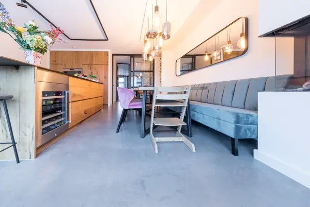Stijlvolle, lichte open woonruimte met moderne houten keuken, grijsblauwe loungebank en designverlichting. De naadloze vloer zorgt voor een schoon, samenhangend geheel.