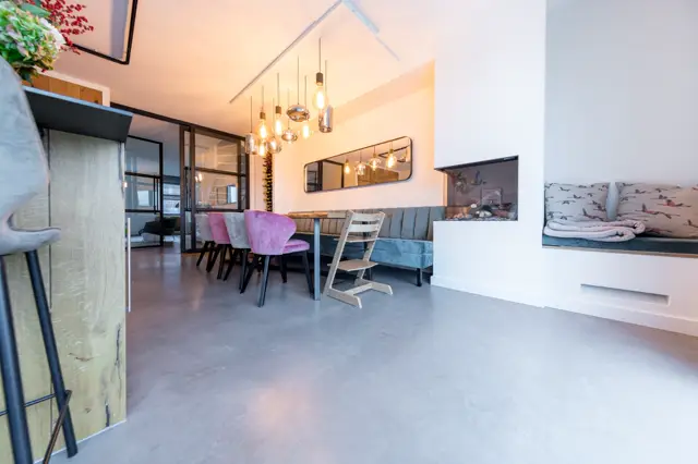 Ruime, lichte woonkamer met geïntegreerde keuken, uitgerust met een strakke naadloze vloer in warm grijs. Minimalistisch design met natuurlijke lichtinval en designer-hanglamp boven de eettafel.