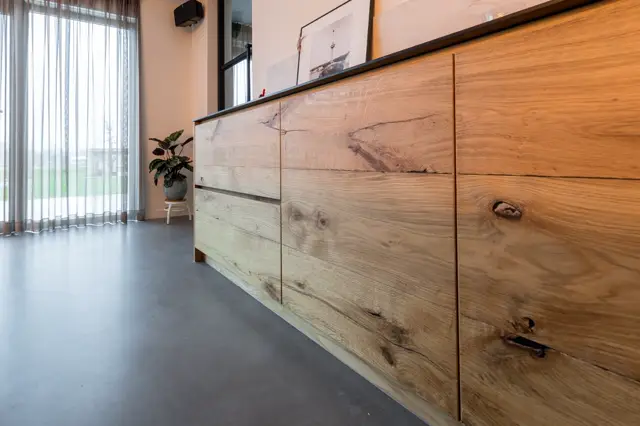Moderne hal met grijze naadloze vloer en houten wandelement met natuurlijke textuur. Helder natuurlijk licht van grote ramen, minimalistische inrichting met plant en artwork.