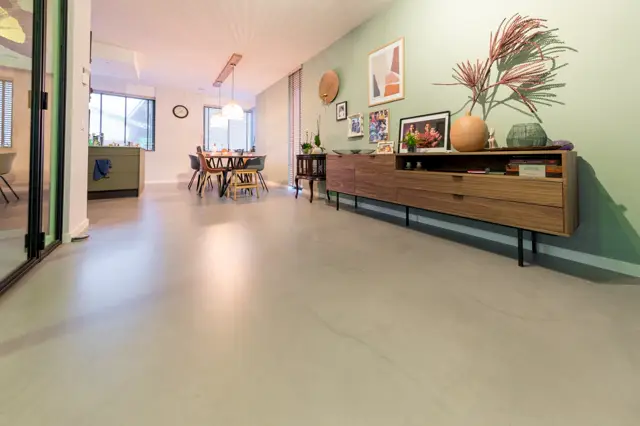 Ruime, lichte woon- en eetkamer met naadloze vloer in warm beige. Strakke architectuur met grote ramen, groen geacccentueerde wanden en minimalistisch ingericht met houten meubels en plantendecoratie.