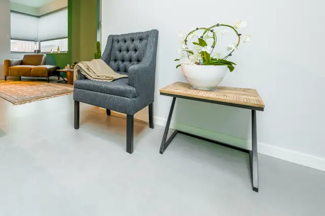Een serene, minimalistisch ingerichte woonhoek met een lichte naadloze vloer die rust en ruimtelijkheid uitstraalt. Moderne meubels in grijs en hout creëren een gebalanceerd, opgeruimd geheel met natuurlijk licht.