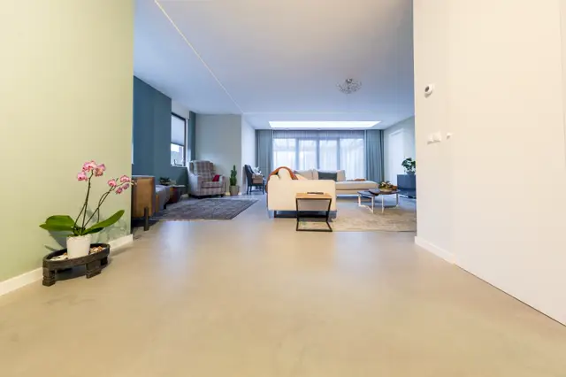 Ruime, licht ingestelde woonkamer met een uniforme naadloze vloer in warm beige. De ruimte ademt luxe uit met een minimalistisch design, grote ramen, verfijnde inrichting en subtiele kleurkeuzes.